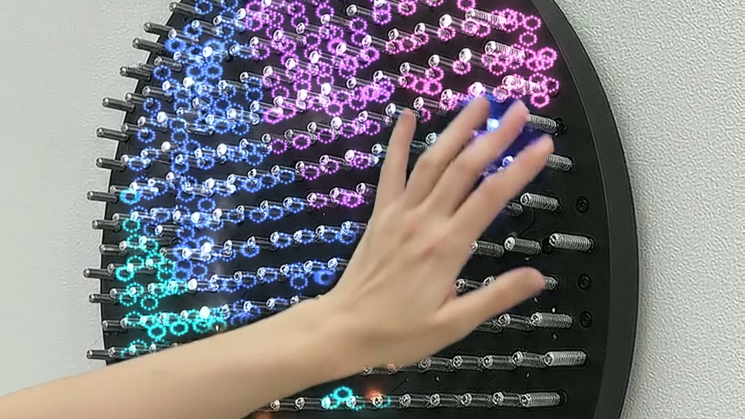 A hand on the Ripple array, the springs around it lit in rings of purple, pink and cyan, the rest still dark